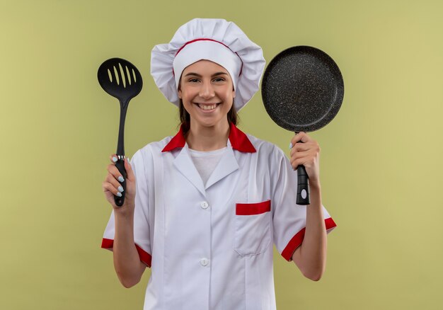 La giovane ragazza caucasica sorridente del cuoco in uniforme dello chef tiene la spatola e la padella isolata su spazio verde con lo spazio della copia