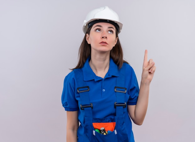 La giovane ragazza confusa del costruttore con il casco di sicurezza bianco e l'uniforme blu indica in su e cerca su fondo bianco isolato con lo spazio della copia