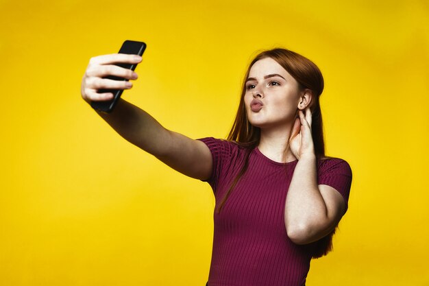 La giovane ragazza della testarossa sta facendo il selfie sul telefono cellulare e sta inviando il bacio dell'aria