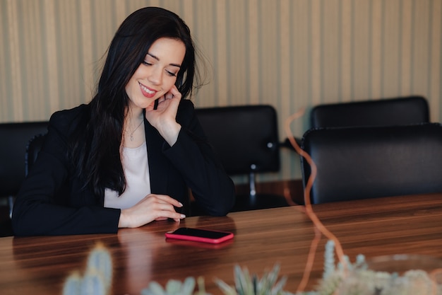 La giovane ragazza emozionale attraente in vestiti di stile di affari si siede allo scrittorio con il telefono in ufficio o pubblico