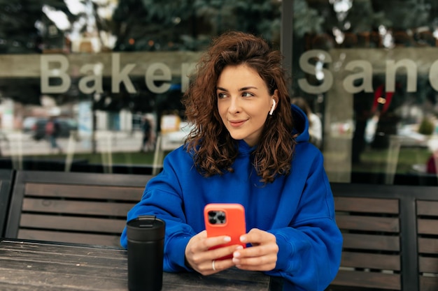 La giovane ragazza europea spensierata sta usando uno smartphone moderno seduto all'aperto con il caffè La bruna riccia indossa pullover e jeans Concetto di tecnologia