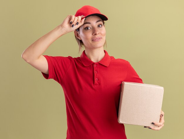 La giovane ragazza graziosa di consegna soddisfatta in uniforme mette la mano sul cappuccio e tiene il cardbox su verde oliva