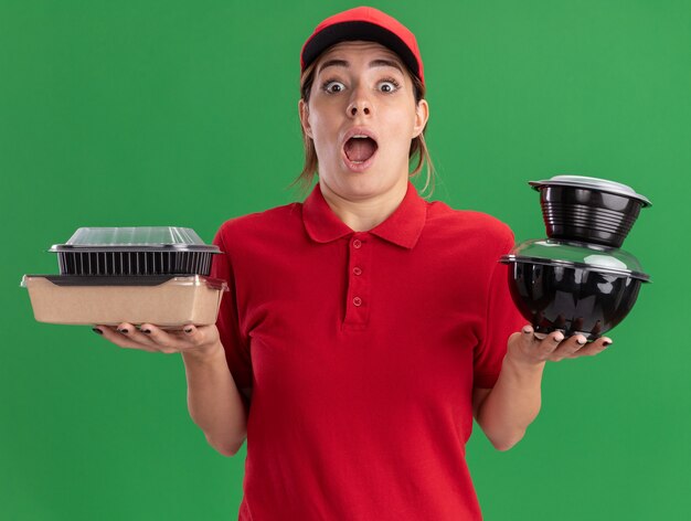 La giovane ragazza graziosa di consegna sorpresa in uniforme tiene i contenitori per alimenti sul verde