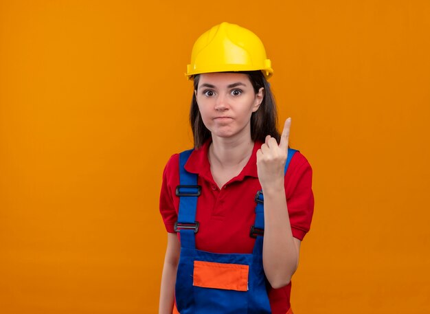 La giovane ragazza infastidita del costruttore mostra un gesto del dito su fondo arancio isolato con lo spazio della copia