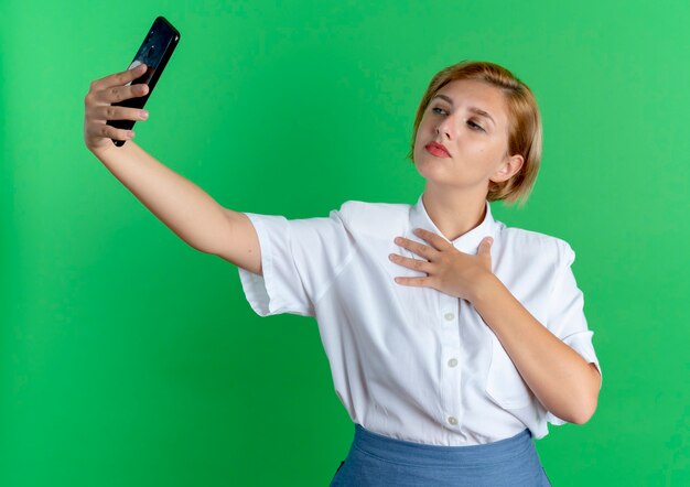 La giovane ragazza russa bionda fiduciosa guarda al telefono prendendo selfie mette la mano sul petto isolato su sfondo verde con spazio di copia