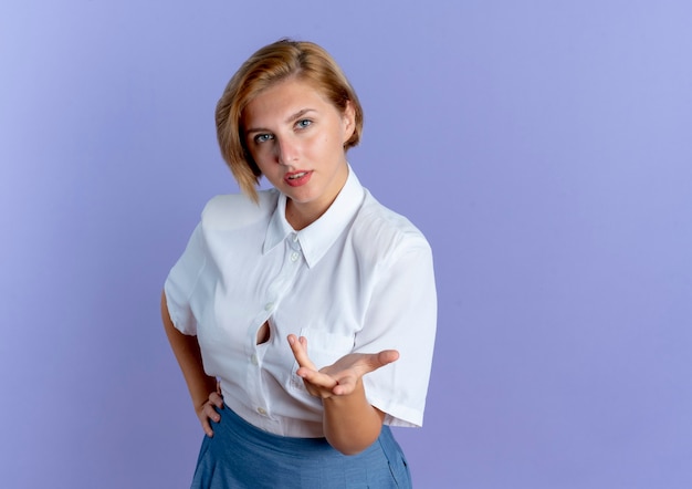 La giovane ragazza russa bionda fiduciosa guarda e punta con la mano alla macchina fotografica isolata su fondo viola con lo spazio della copia