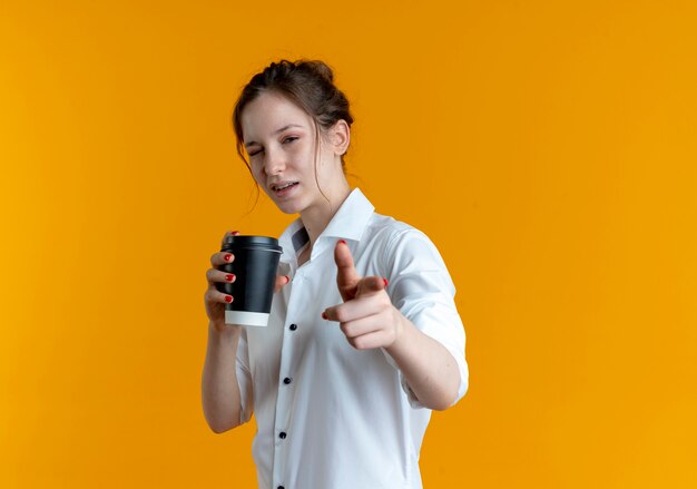 La giovane ragazza russa bionda fiduciosa lampeggia l'occhio che tiene la tazza di caffè e indica la macchina fotografica isolata sullo spazio arancione con lo spazio della copia