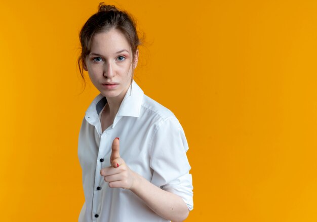 La giovane ragazza russa bionda fiduciosa osserva e indica la macchina fotografica isolata sullo spazio arancione con lo spazio della copia