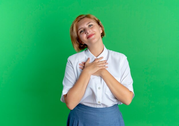 La giovane ragazza russa bionda lieta mette le mani sul petto che guarda l'obbiettivo isolato su priorità bassa verde con lo spazio della copia