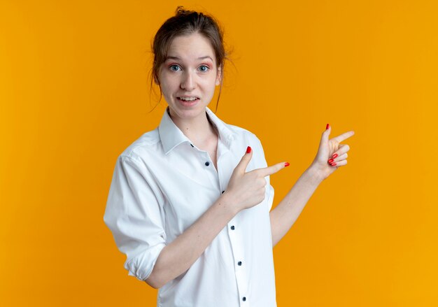 La giovane ragazza russa bionda sorpresa indica a lato con due mani isolate sullo spazio arancione con lo spazio della copia