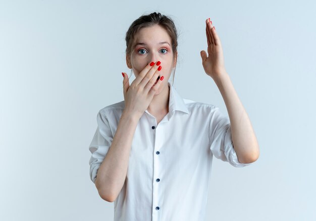 La giovane ragazza russa bionda sorpresa mette la mano sulla bocca e alza la mano isolata su uno spazio bianco con lo spazio della copia