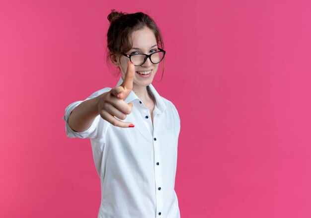 La giovane ragazza russa bionda sorridente con gli occhiali punta alla macchina fotografica isolata sullo spazio rosa con lo spazio della copia