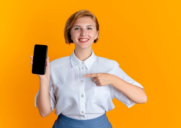 La giovane ragazza russa bionda sorridente indica al telefono isolato su priorità bassa arancione con lo spazio della copia