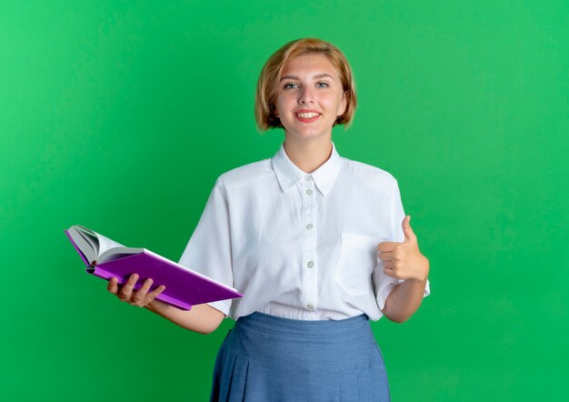 La giovane ragazza russa bionda sorridente thumbs up e tiene il libro isolato su priorità bassa verde con lo spazio della copia