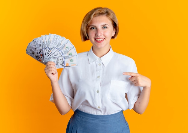 La giovane ragazza russa bionda sorridente tiene e indica i soldi isolati su fondo arancio con lo spazio della copia