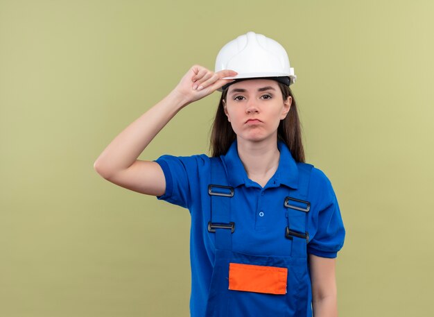 La giovane ragazza sicura del costruttore con il casco di sicurezza bianco e l'uniforme blu tiene il hemlet e guarda la macchina fotografica su fondo verde isolato