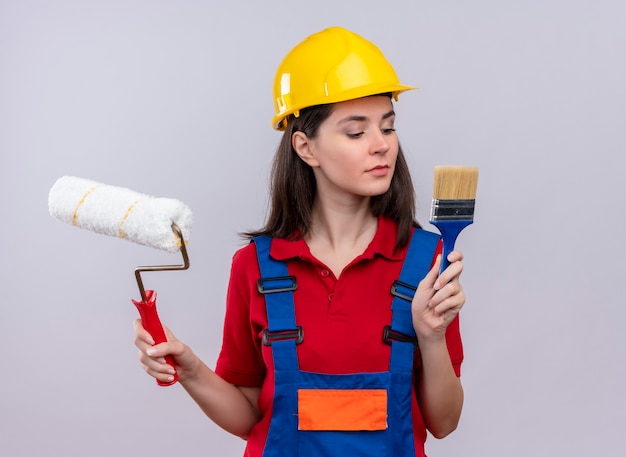 La giovane ragazza sicura del costruttore tiene il rullo di vernice e il pennello su fondo bianco isolato