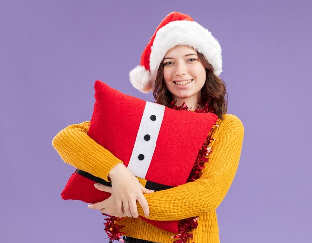 La giovane ragazza slava sorridente con il cappello della Santa e con la ghirlanda intorno al collo tiene il cuscino decorato isolato su fondo viola con lo spazio della copia