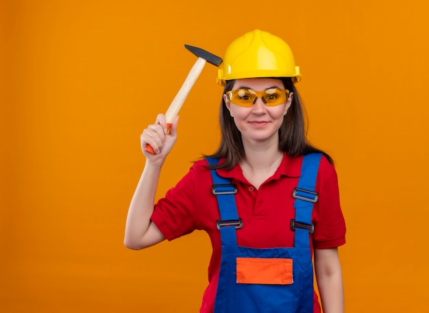 La giovane ragazza sorridente del costruttore con gli occhiali di sicurezza tiene il martello su fondo arancio isolato con lo spazio della copia