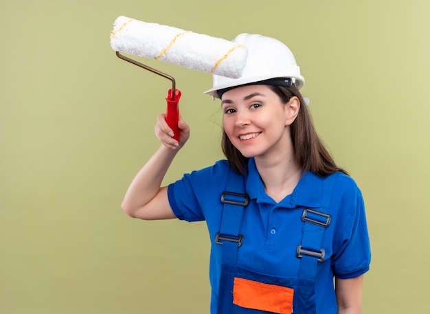 La giovane ragazza sorridente del costruttore con il casco di sicurezza bianco e l'uniforme blu tiene il rullo di vernice su fondo verde isolato