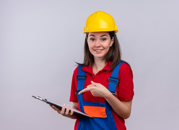 La giovane ragazza sorridente del costruttore tiene la lavagna per appunti e punti alla lavagna per appunti su fondo bianco isolato con lo spazio della copia