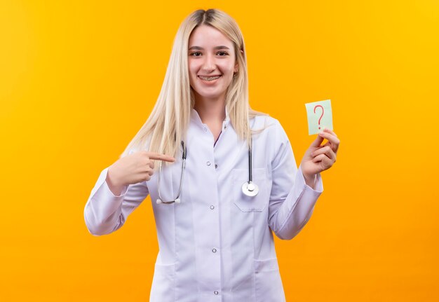 La giovane ragazza sorridente del medico che porta lo stetoscopio in camice medico e il tutore dentale indica il punto interrogativo di carta sulla sua mano su fondo giallo isolato