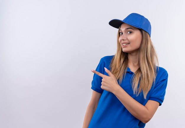 La giovane ragazza sorridente di consegna che porta l'uniforme blu e le punte del cappuccio al lato isolato sulla parete bianca con lo spazio della copia