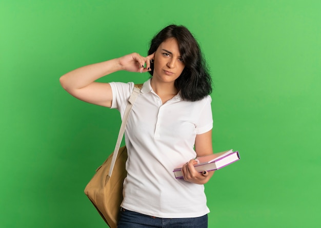 La giovane scolara abbastanza caucasica infastidita che porta la borsa posteriore chiude l'orecchio che tiene i libri sul verde con lo spazio della copia