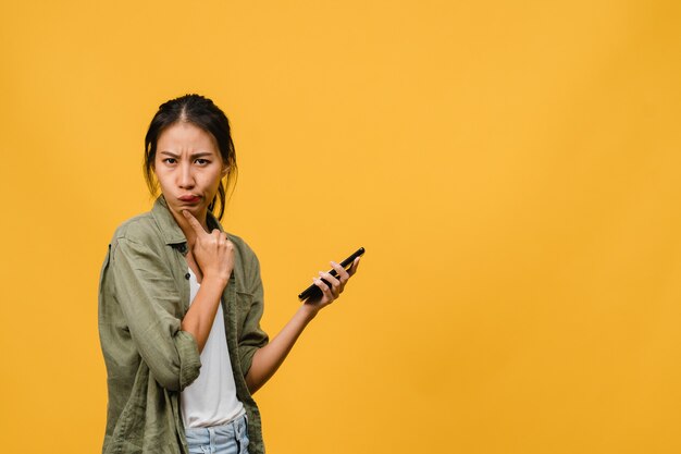 La giovane signora asiatica che usa il telefono con un'espressione positiva si è vestita con un panno casual sul muro giallo