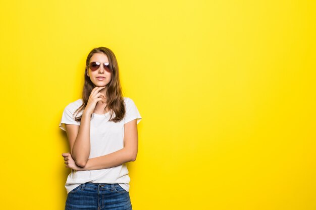 La giovane signora di pensiero in maglietta bianca e jeans blu rimane davanti al fondo giallo dello studio