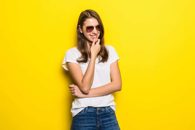 La giovane signora di pensiero in maglietta bianca e jeans blu rimane davanti al fondo giallo dello studio