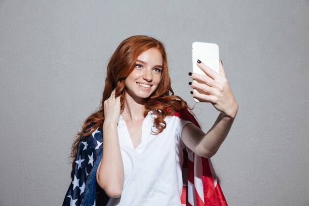 La giovane signora felice della testarossa con la bandiera di USA fa il selfie
