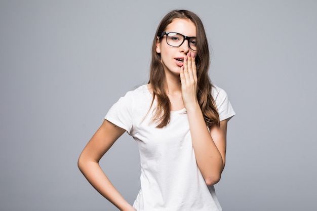 La giovane signora graziosa timida in vetri in maglietta bianca e blue jeans sta davanti al fondo bianco dello studio