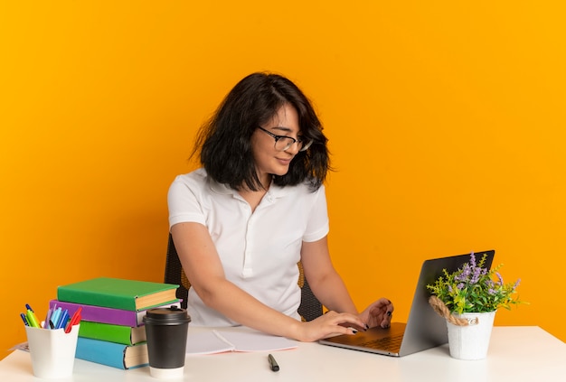 La giovane studentessa abbastanza caucasica con gli occhiali si siede allo scrittorio con i lavori degli strumenti della scuola e guarda il computer portatile isolato sullo spazio arancione con lo spazio della copia