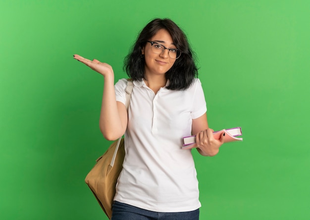 La giovane studentessa abbastanza caucasica confusa con gli occhiali e la borsa posteriore tiene i libri e solleva la mano sul verde con lo spazio della copia