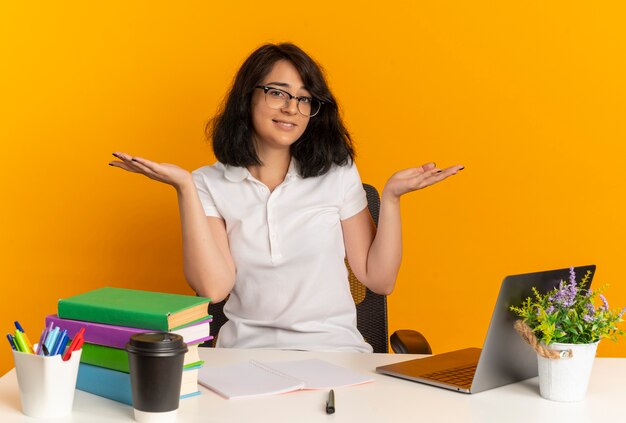 La giovane studentessa abbastanza caucasica confusa con gli occhiali si siede allo scrittorio con gli strumenti della scuola tiene le mani aperte isolate sullo spazio arancione con lo spazio della copia