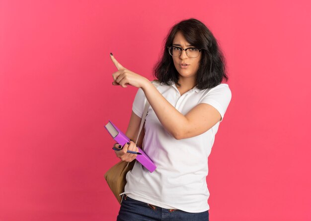 La giovane studentessa abbastanza caucasica infastidita con gli occhiali e la borsa posteriore tiene la penna e il libro che punta a lato sul colore rosa con lo spazio della copia