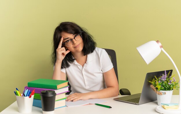 La giovane studentessa abbastanza caucasica infastidita con gli occhiali si siede allo scrittorio con gli strumenti della scuola mette il dito sulla testa isolata su spazio verde con lo spazio della copia