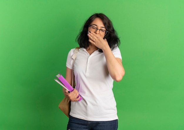 La giovane studentessa abbastanza caucasica sonnolenta con gli occhiali e la borsa posteriore mette la mano sulla bocca che tiene i libri sul verde con lo spazio della copia