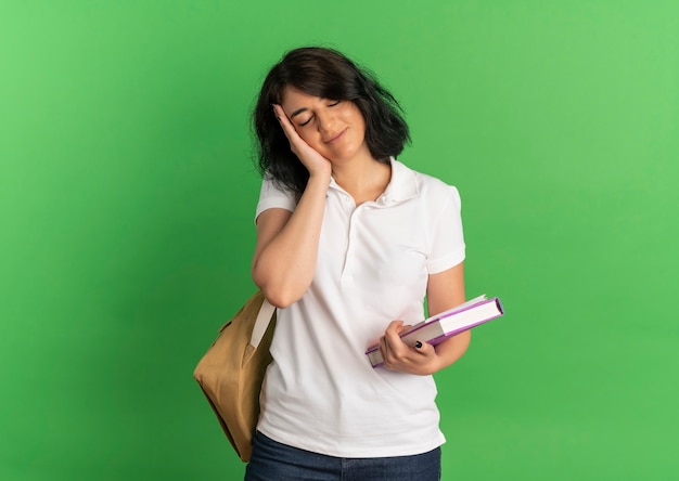 La giovane studentessa graziosa caucasica soddisfatta che porta la borsa posteriore mette la mano sulla faccia che tiene i libri sul verde con lo spazio della copia