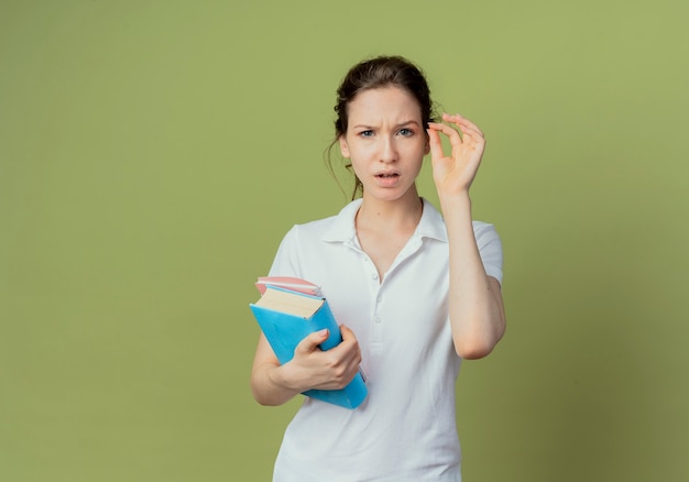 La giovane studentessa graziosa confusa che tiene il libro e il blocco note che tengono la mano nell'aria finge che tiene qualcosa isolato su fondo verde oliva con lo spazio della copia