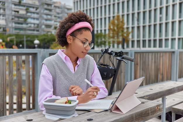 La giovane studentessa guarda il webinar o il video tutorial concentrato sullo schermo del tablet scrive appunti nelle note del blocco note le informazioni sul progetto si pongono all'esterno contro un'atmosfera accogliente crea un piano