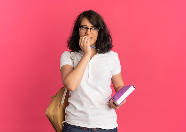 La giovane studentessa piuttosto caucasica ansiosa con gli occhiali e la borsa posteriore morde le dita che tengono la penna e il libro sul rosa con lo spazio della copia