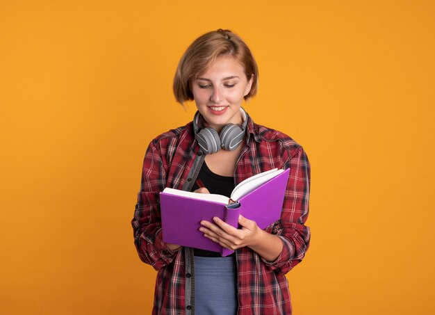 La giovane studentessa slava sorridente con le cuffie che indossa lo zaino scrive sul libro con la penna