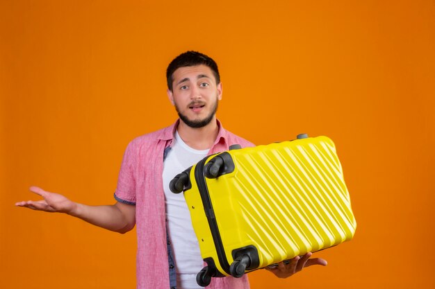 La giovane valigia bella della tenuta del tipo del viaggiatore incapace e confusa che esamina la macchina fotografica che sta con il braccio si è alzata avendo dubbi sopra fondo arancio