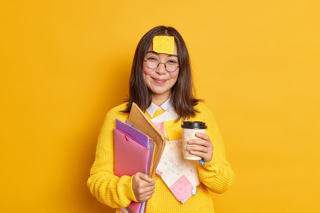 La lavoratrice asiatica positiva tiene la tazza di caffè usa e getta tiene le cartelle ha un adesivo con il grafico bloccato sulla fronte si è rotto dopo l'apprendimento dell'esame.