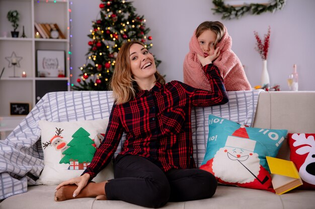 La madre sorridente che si siede sul divano tiene le guance delle figlie avvolte in una coperta che gode del tempo di Natale a casa