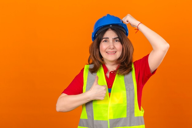 La maglia d'uso della costruzione dell'ingegnere della donna e il casco di sicurezza sorpresi con la mano sulla testa per l'errore ricordano l'errore che mostra il pollice su che sembra confuso sopra la parete arancio isolata