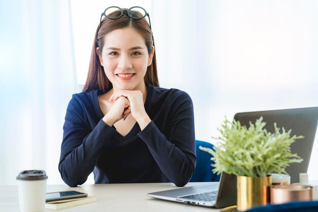 La maglietta nera della donna di affari asiatica casuale astuta creativa lavora a casa con lo smartphone e il computer portatile con la luce del mattino dalla finestra