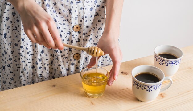 La mano della donna che gocciola il miele in vetro con la tazza delle tazze di tè sullo scrittorio di legno